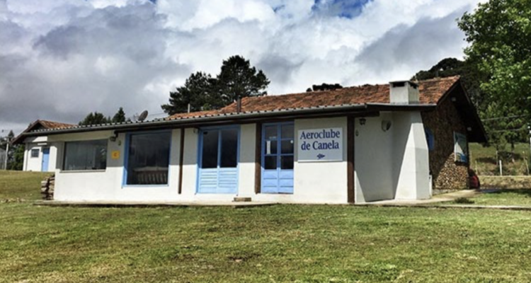 Sede do Aeroclube de Canela, construção branca com portas azuis cercada por gramado e pinheiros da Serra Gaúcha